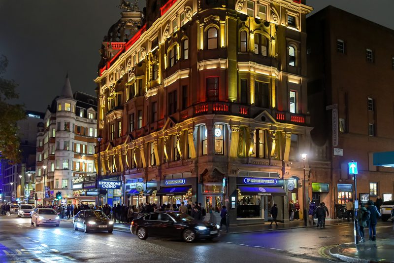 Hippodrome Casino na rogu Cranbourn Street w Londynie, oświetlone wieczorem, obok przejeżdżają samochody